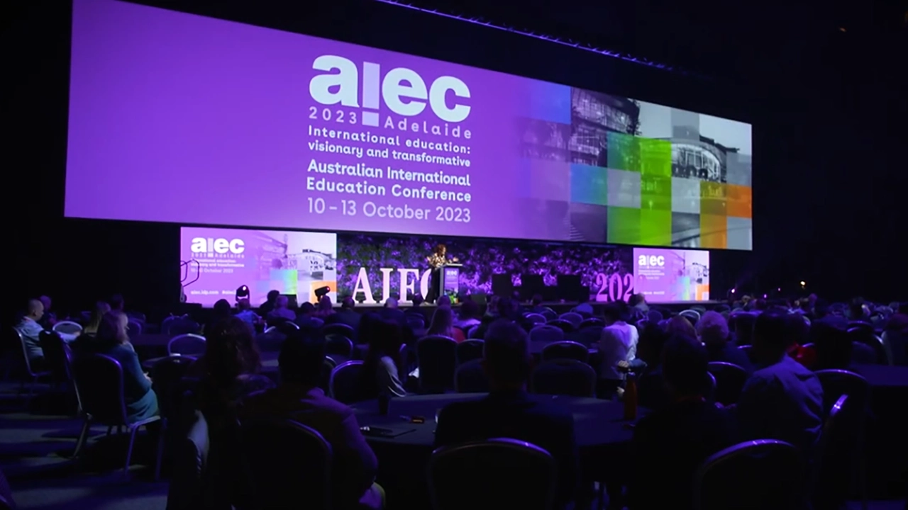 Purple illuminated banner on a stage with a woman at a lecturn speaking in a theatre with a seated crowd looking to the stage