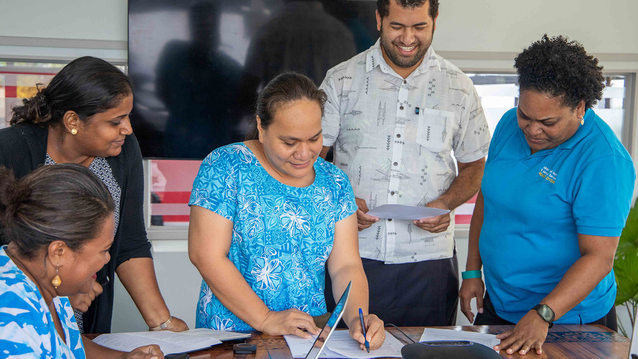 Group of people from the Fiji Facility standing around a table smiling and looking at some documents