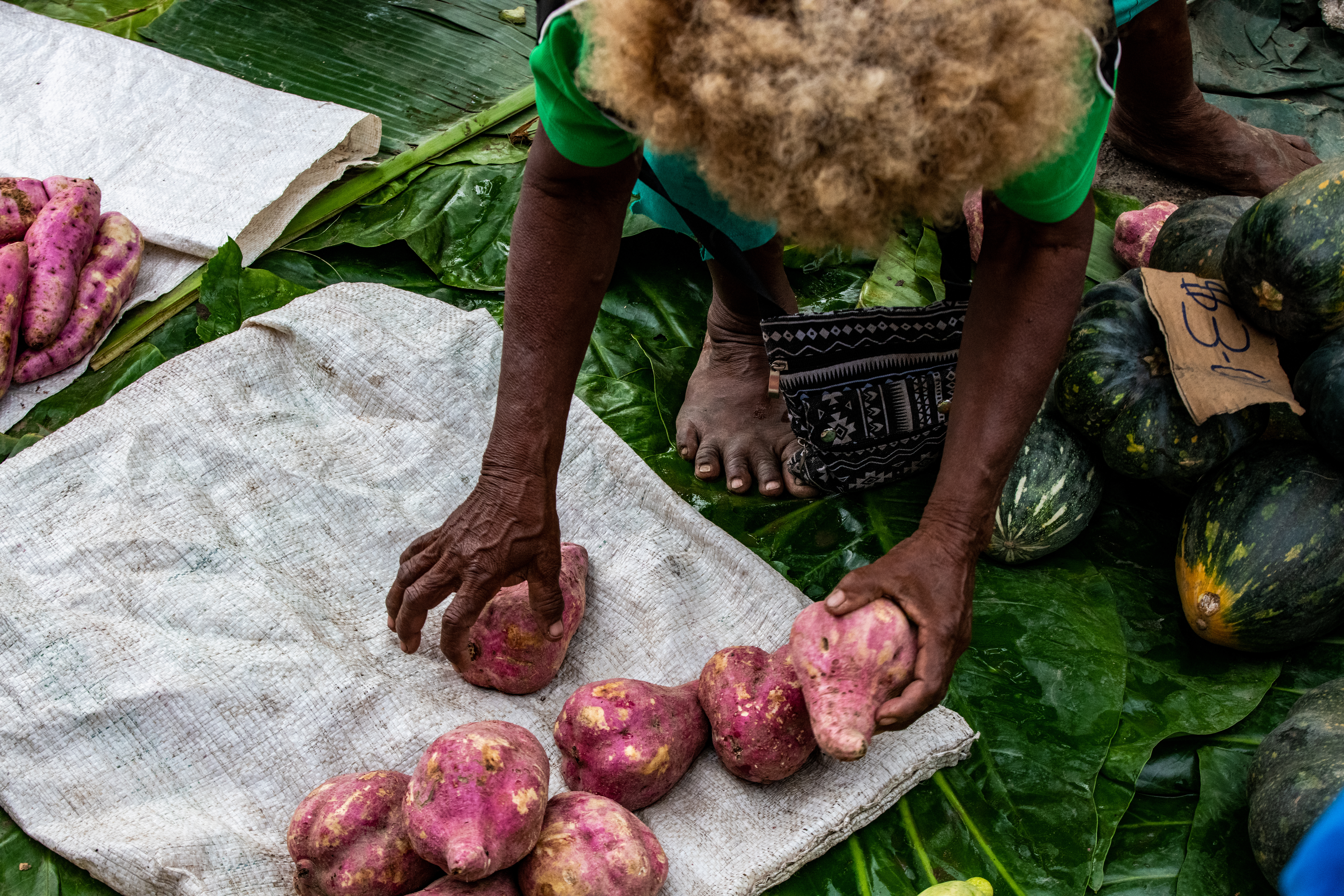 Aerial view of a person crouching over a bag on the ground with potatoes on the bag. There are also other vegetable products on the ground. The setting is at a market in Honiara, the Solomon Islands